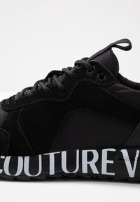 Svart sneaker med en design i mocka och tyg, med en texturerad sula och "COUTURE" tryckt i ljusblått på sidan.