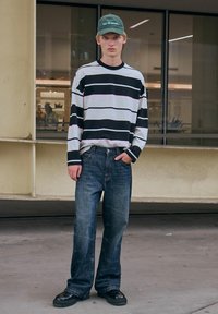 Camicia a maniche lunghe a righe bianca e nera, abbinata a jeans a gamba larga blu scuro e scarpe nere con lacci. Berretto verde con testo.
