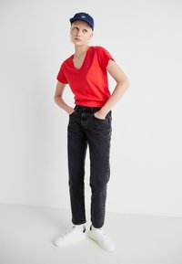 T-shirt rossa a scollo a V, jeans neri a vestibilità slim e sneakers bianche. Gli accessori includono un cappellino blu e orecchini a cerchio. Design e colori semplici.