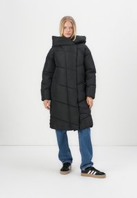 Melns pūkains mētelis ar kapuci, kam ir quilted dizains. Tas ir līdz ceļiem un apvienots ar zilām biksēm un melniem sporta apaviem.