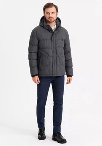 Grå polstret jakke med hætte, der har sidelommer og en lynlås foran, båret over mørke denimjeans og sorte støvler.