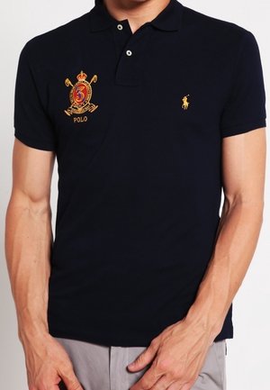 Granatowa koszulka polo z bawełny, z haftowanym złotym logo na klatce piersiowej, krótkimi rękawami i kołnierzykiem zapinanym na dwa guziki.