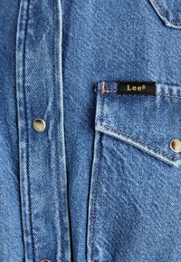 Gaiši zila džinsa krekls ar klasisku dizainu, viena krūšu kabata, sudraba pogas ar snap sistēmu un melna etiķete ar dzeltenu tekstu.