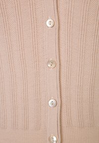 Cardigan en maille beige clair avec une texture en côtes verticales, comportant six boutons ronds en perles le long de l'avant. Matériau doux.