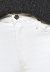 Gros plan sur la taille et le zip d'un jean en denim blanc avec un bouton en cuivre sous un pull en mailles gris foncé.