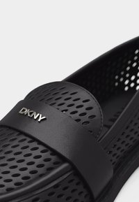 Chaussure noire perforée à enfiler avec une large bande sur le dessus portant le logo DKNY argenté.