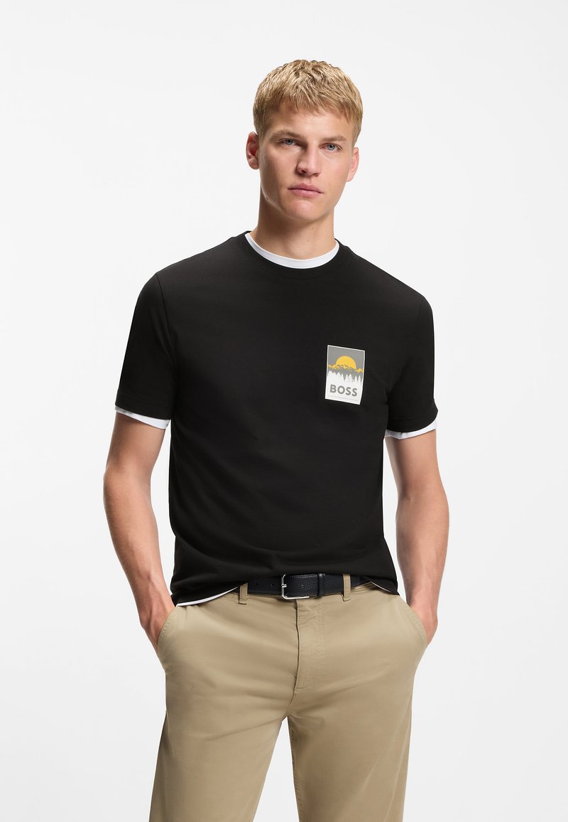 Νεαρός άνδρας που φοράει μαύρο T-shirt με κίτρινο ηλιοβασίλεμα και λογότυπο "BOSS", μπεζ παντελόνι, με τα χέρια στις τσέπες, στέκεται μπροστά σε λευκό φόντο.