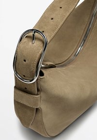 Blød taupe ruskinds håndtaske med en sølvmetal spænde på en bred rem. Har en lynlåslukning og en glat tekstur, ideel til hverdagsbrug.