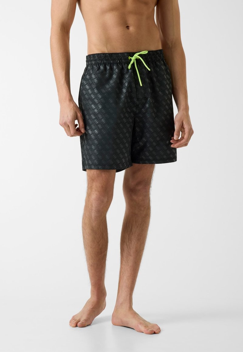 Torse et jambes d'un homme portant un short de bain noir à motifs avec un cordon vert néon, debout pieds nus sur une surface claire.