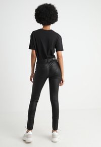 Person with curly hair wearing a black t-shirt, black leather pants, and white sneakers, standing facing a plain white background.