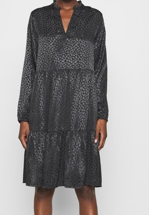 Mujer con un vestido negro de manga larga con un sutil estampado de leopardo, falda de volantes en varias capas y cuello con volantes, posando contra un fondo liso.