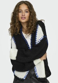 Cárdigan extragrande en blanco y negro con costuras azules. Textura de punto grueso, que presenta audaces patrones diagonales y mangas anchas.