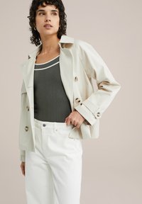 Ljusbeige, dubbelknäppt trenchcoat med stora knappar. Bärs över en mörk tanktop och vita vida jeans. Slätt tyg, skräddarsydd passform.