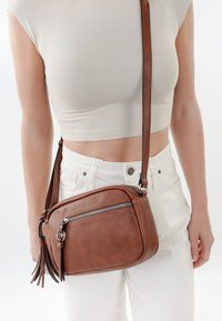 Sieviete, kas valkā bēša krāsas īso topu un baltas bikses, nēsā brūnu ādas somiņu ar priekšējā rāvējslēdzēja kabatu un bārkstīm.