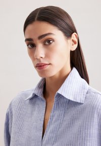 Camicia a righe blu chiaro con colletto morbido e pattina con bottoni, realizzata in un tessuto leggero, con una sottile texture e dettagli di pieghe.