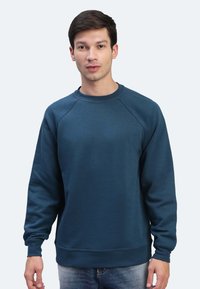 Felpa di un verde scuro con maniche lunghe, colletto a girocollo a coste e polsini a coste. Tessuto morbido con vestibilità rilassata, indossata sopra jeans in denim.