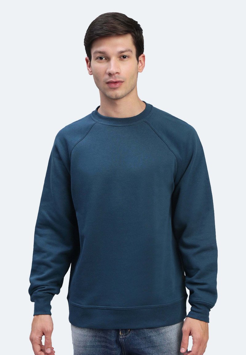 Felpa di un verde scuro con maniche lunghe, colletto a girocollo a coste e polsini a coste. Tessuto morbido con vestibilità rilassata, indossata sopra jeans in denim.