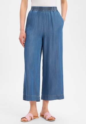 Femme portant un pantalon en jean bleu à jambes larges avec taille élastique et sandales plates ouvertes roses sur fond blanc.