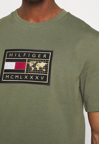 Camiseta de algodón color verde oliva con un gran parche bordado negro, con acentos en rojo, blanco y dorado, que muestra el nombre y el logo de la marca.