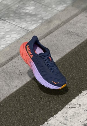 Running shoe with a navy mesh upper, pink and purple gradient midsole, orange accents, and textured rubber sole, set on textured surface.