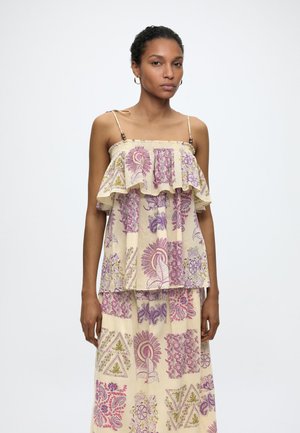 Femme portant une robe beige sans manches avec des motifs floraux et géométriques violets et roses, debout devant un fond blanc uni.