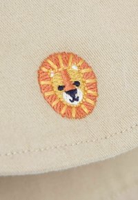 Beige szövet kerek hímzett mintával, amely egy oroszlánarcot ábrázol, narancssárga, sárga és fekete árnyalatokkal díszítve.