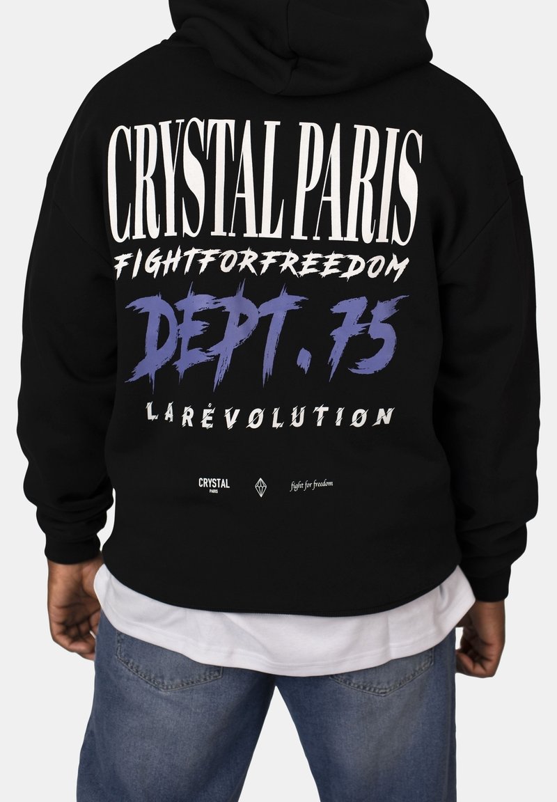 Mustapaita, jossa on suuri valkoinen ja purppura teksti selkäpuolella, sisältäen grafiikoita kuten "CRYSTAL PARIS" ja "DEPT. 75." Puuvillamateriaalia.