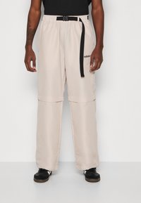 Světlé béžové sportovní kalhoty s černým pásem, které mají volný střih, kapsy na zip a vyšitým logem na levé noze.