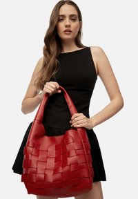 Mujer vestida con un vestido negro sin mangas, sosteniendo un gran bolso de cuero trenzado rojo por sus asas, frente a un fondo liso.