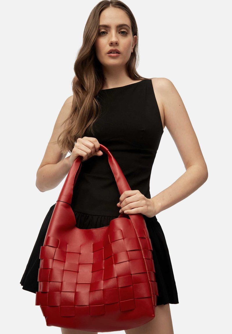 Mujer vestida con un vestido negro sin mangas, sosteniendo un gran bolso de cuero trenzado rojo por sus asas, frente a un fondo liso.