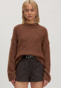 Frau mit schulterlangem blonden Haar trägt einen braunen flauschigen Pullover, einen schwarzen Gürtel, dunkle strukturierte Shorts und goldene Creolen vor einem schlichten Hintergrund.