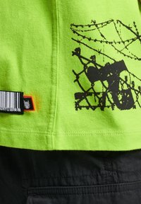 Svěží zelené bavlněné tričko má černý grafický design, štítek s čárovými kódy a oranžový akcent na lemu.