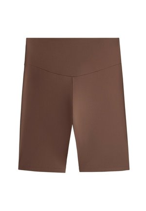 Kvinde iført en hvid fleecetrøje over en brun- og hvidstribet top, brune cykelshorts, brune sokker og brune sneakers, stående på en grå baggrund.