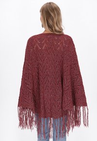 Poncho en tricot bourgogne avec un motif chevrons ouvert, présentant des bords frangés et un design ample et fluide. Texture douce et matériau léger.