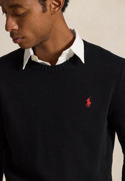 Joven mirando hacia abajo con un jersey negro de punto sobre una camisa blanca con cuello y un pequeño logo rojo bordado en el pecho.