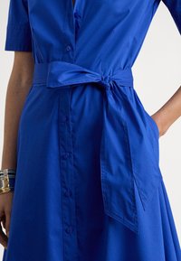 Modra bombažna srajčna obleka z opasanim pasom, kratkimi rokavi in sprednjim zapenjanjem na gumbe. Ima razširjen spodnji rob in podrobnost stranskega žepa.