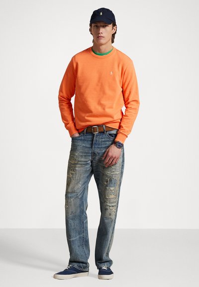 Orange sweatshirt with a round neck, worn over a green shirt; paired with distressed blue jeans and navy canvas shoes. Accessories include a navy cap and a watch.