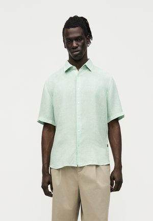 Homme avec des dreadlocks portant une chemise verte claire à manches courtes boutonnée et un pantalon beige, debout devant un fond blanc uni.