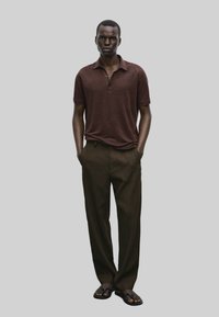 Polo shirt corto marrone con maniche corte abbinato a pantaloni eleganti marroni scuri e ciabatte nere. Il modello sta in piedi con le mani in tasca.