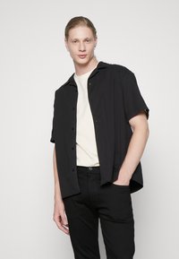 Schwarzes kurzärmliges Hemd mit Knopfverschluss, getragen über einem hellbeigen T-Shirt, kombiniert mit schwarzen Hosen. Glatte Textur, klassischer Schnitt.