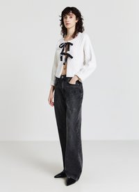 Weißes gesteppte Cropped-Jackett mit schwarzem Bandverschluss, kombiniert mit hoch taillierten schwarzen Denim-Weitbein-Hosen und spitzen schwarzen Schuhen.