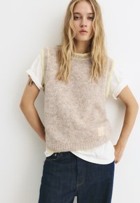 Hellbrauner Strickweste mit limegrüner Einfassung, texturiertem Stoff und einem quadratischen Logo-Patch am Saum. Getragen über einem weißen T-Shirt.