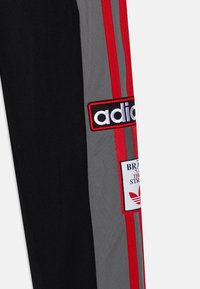 Černé a šedé sportovní kalhoty s červenými pruhy, s nášivkou s textem a logem "adidas" vpředu, vyrobené z texturované tkaniny.