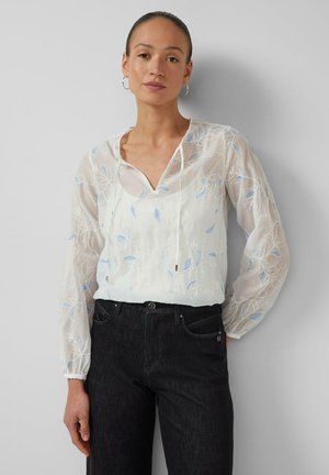 Frau trägt eine durchsichtige weiße Bluse mit blauer Blumenstickerei und schwarze, hoch taillierte Jeans, steht vor einem schlichten hellen Hintergrund.