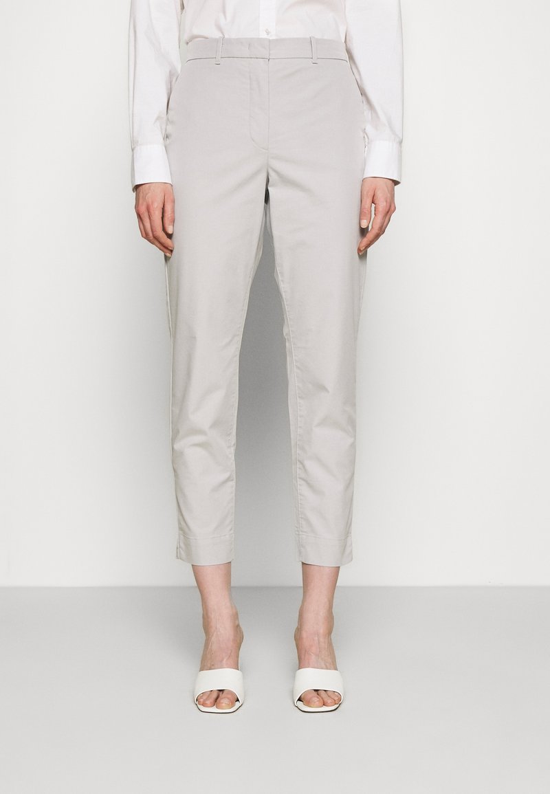 Personne portant un pantalon court gris clair, une chemise blanche boutonnée et des talons ouverts blancs, debout devant un fond uni.