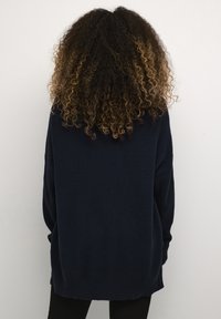 Isik, kellel on lokkis pruunid ja blondid juuksed, seljas tumesinine soonikuga kampsun, näoga selja poole lihtsal heledal taustal.