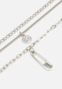 Svetlo srebrna ogrlica z več plasti, ki vključuje različne stile verig, okroglo obesek s profilom in čar na varnostni zatič.