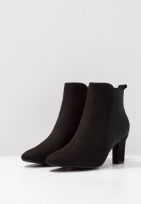 Bottines noires en daim doux avec un bout pointu, des panneaux latéraux élastiques et un talon bloc de 5 cm. Design épuré avec des coutures simples.
