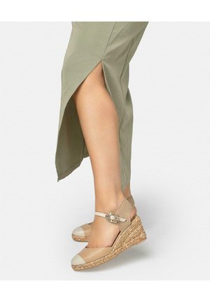 Femme portant des espadrilles compensées beiges à brides autour de la cheville et une jupe verte claire fendue sur le côté, dévoilant la jambe inférieure et le pied.