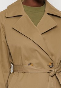 Khaki trencz wytworzony z gładkiej tkaniny, z podwójnym zapięciem na duże, okrągłe guziki oraz związanym paskiem w talii.